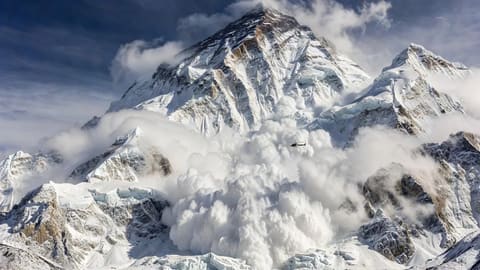 A massive avalanche cascading down a towering Himalayan mountain peak with a small helicopter flying near the enormous cloud of snow, dramatic photorealistic wide-angle shot.