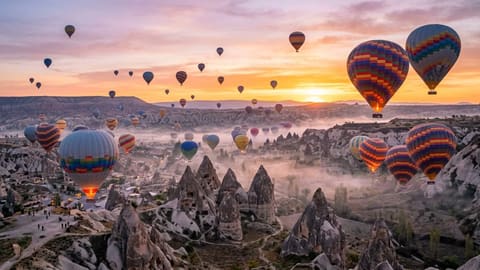 Enormous hot air balloons in vibrant patterns floating over Cappadocia's fairy chimneys at sunrise. Pink and orange sky gradient, mist in the valleys below. Travel landscape, dreamlike atmosphere.