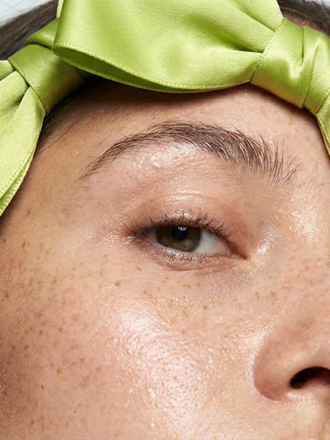 Extreme macro close-up of a young woman's face from nose to forehead. Dewy skin, natural freckles, a single oversized lime-green satin bow perched on her head. Soft studio lighting, beauty editorial, sharp focus on skin texture.