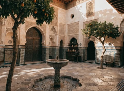 A sunlit Moroccan riad courtyard with intricate zellige tilework, carved stucco arches, a mosaic fountain, and orange trees, warm vintage tones.